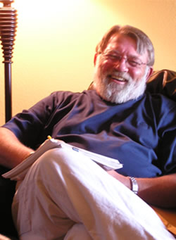 Clay Reynolds in the glow of a lamp laughing. He is wearing a blue shirt and khaki pants. He wears glasses and has a full white beard.